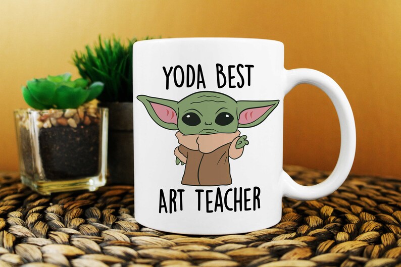 Puede incluir: Taza de cer&aacute;mica blanca con un personaje de dibujos animados verde con orejas grandes y el texto "YODA BEST ART TEACHER" impreso en ella.
