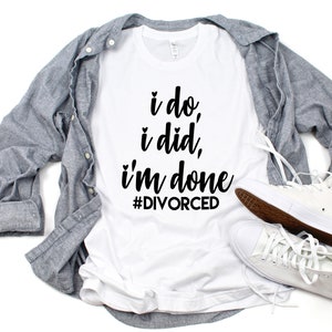 May include: White t-shirt with black text that reads "i do, i did, i'm done #DIVORCED". The shirt is laying on a white surface with a gray button-down shirt and white high-top sneakers.