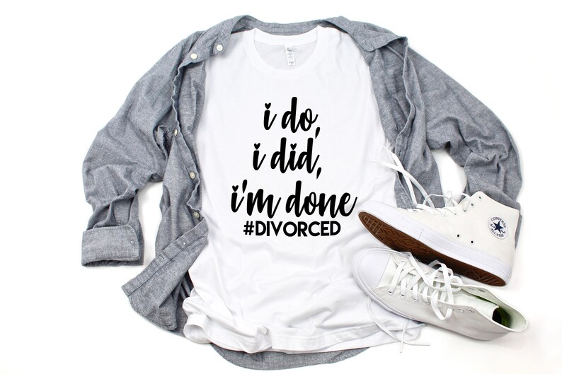 May include: White t-shirt with black text that reads "i do, i did, i'm done #DIVORCED". The shirt is laying on a white surface with a gray button-down shirt and white high-top sneakers.