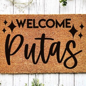 May include: A brown coir doormat with the text "WELCOME PUTAS" in black lettering. The text is surrounded by small black star shapes.