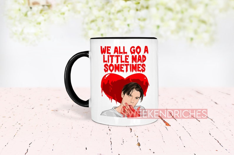 May include: White ceramic coffee mug with a black handle. The mug has a red heart with a dripping blood effect and a cartoon image of a man with a bloody hand. The text on the mug reads "We all go a little mad sometimes".