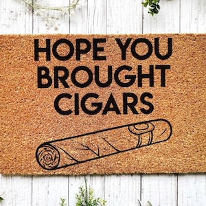 K&ouml;nnte beinhalten: Eine braune Kokosfaser-Fu&szlig;matte mit dem Text "HOPE YOU BROUGHT CIGARS" in schwarzen Buchstaben. Unter dem Text befindet sich eine Schwarzwei&szlig;illustration einer Zigarre.