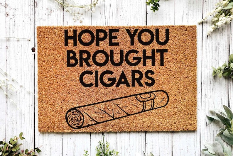 K&ouml;nnte beinhalten: Eine braune Kokosfaser-Fu&szlig;matte mit dem Text "HOPE YOU BROUGHT CIGARS" in schwarzen Buchstaben. Unter dem Text befindet sich eine Schwarzwei&szlig;illustration einer Zigarre.