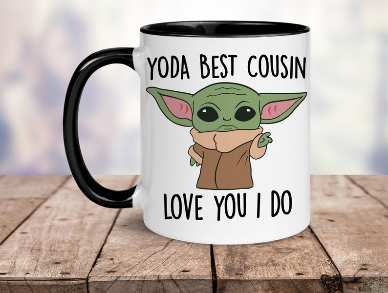 Puede incluir: Taza de cer&aacute;mica blanca con borde y asa negros. La taza presenta una imagen de dibujos animados de un personaje alien&iacute;gena verde con ojos grandes y una t&uacute;nica marr&oacute;n. El texto "YODA BEST COUSIN LOVE YOU I DO" est&aacute; impreso en la taza.