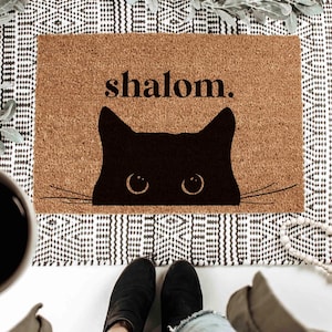 Könnte beinhalten: Eine braune Kokosmatte mit dem Wort "shalom." in Schwarz gedruckt. Eine schwarze Katzensilhouette mit weißen Augen lugt hinter dem Text hervor.