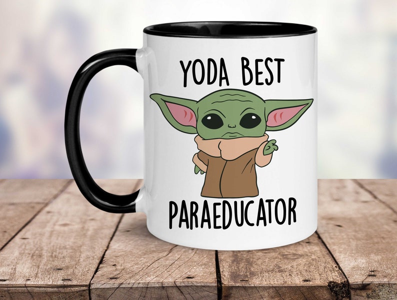 K&ouml;nnte beinhalten: Wei&szlig;e Keramik-Kaffeetasse mit schwarzem Rand und Henkel. Die Tasse zeigt ein Cartoon-Bild eines gr&uuml;nen Au&szlig;erirdischen mit gro&szlig;en Ohren und einem braunen Mantel. Der Text "YODA BEST PARAEDUCATOR" ist auf die Tasse gedruckt.