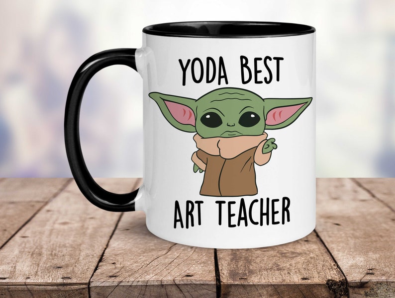 Puede incluir: Taza de caf&eacute; de cer&aacute;mica blanca con borde y asa negros. La taza presenta una imagen de dibujos animados de un personaje alien&iacute;gena verde con orejas grandes y el texto "Yoda Best Art Teacher".