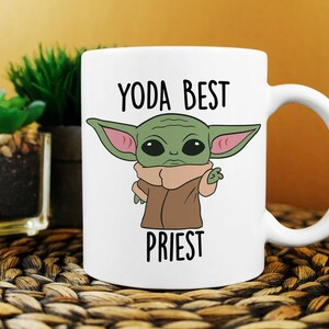 Puede incluir: Taza de cer&aacute;mica blanca con un personaje de dibujos animados verde con orejas grandes y el texto "YODA BEST PRIEST" impreso en ella.