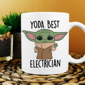 May include: White ceramic mug featuring a cartoon image of a green alien character with large ears and the text "YODA BEST ELECTRICIAN" in black. The mug has a curved handle and is set against a blurred background.