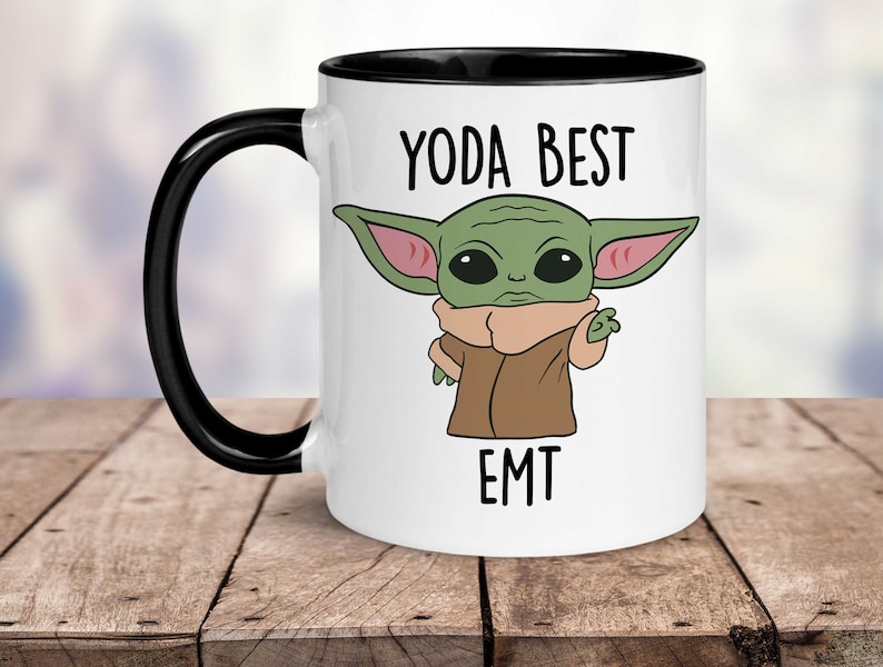 Op de afbeelding: Witte keramische koffiemok met een zwarte rand en handvat. De mok heeft een cartoonafbeelding van een groen buitenaards wezen met grote oren en de tekst "YODA BEST EMT".