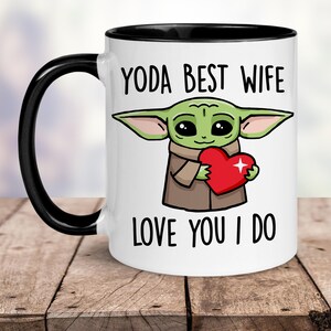 Puede incluir: Taza de cer&aacute;mica blanca con borde y asa negros. La taza presenta una imagen de dibujos animados de un extraterrestre verde sosteniendo un coraz&oacute;n rojo con el texto "YODA BEST WIFE LOVE YOU I DO".