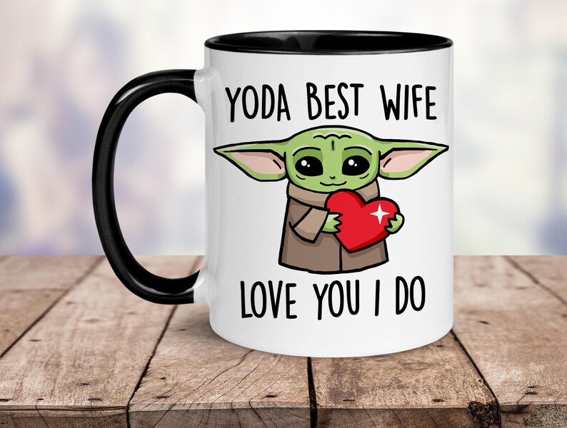 Puede incluir: Taza de cer&aacute;mica blanca con borde y asa negros. La taza presenta una imagen de dibujos animados de un extraterrestre verde sosteniendo un coraz&oacute;n rojo con el texto "YODA BEST WIFE LOVE YOU I DO".