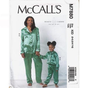 May include: McCall's M7880 sewing pattern envelope featuring a green satin pajama set for adults and children. The adult set includes a button-down shirt with a collar and long pants. The child's set features a button-down top and matching pants.