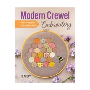 May include: A book titled "Modern Crewel Embroidery" with a stitched sampler in a wooden hoop. The sampler features a honeycomb design with various colors and patterns. A cartoon bee is stitched on the right side. Purple flowers are scattered around the book.