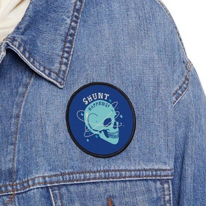 May include: A blue denim jacket with a round patch featuring a skull with a blue and green design. The patch has the text "Shunt Happens" around the skull.