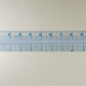 May include: A white glue stick measuring 10 inches in length, placed on a ruler with blue markings.