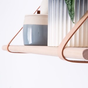 May include: A wooden shelf with leather straps hangs from the ceiling. Two ceramic containers, one blue and white and the other white with a blue ombre effect, sit on the shelf. A small green plant is in the white container.
