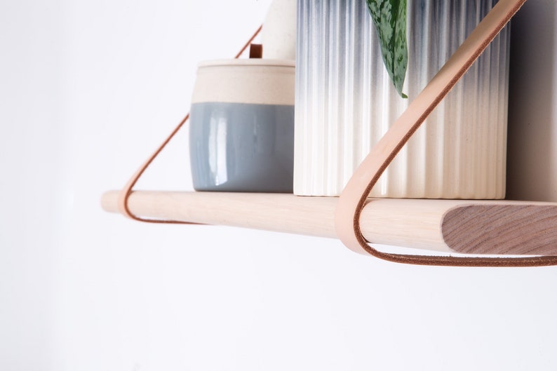 May include: A wooden shelf with leather straps hangs from the ceiling. Two ceramic containers, one blue and white and the other white with a blue ombre effect, sit on the shelf. A small green plant is in the white container.