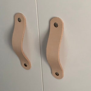 Pode incluir: Duas puxadores de armário de couro marrom claro com detalhes metálicos prateados. Os puxadores estão fixados a uma porta de armário branca.