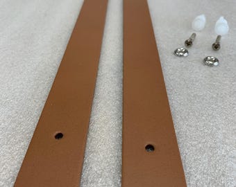 May include: Two long, brown leather straps with rounded ends and pre-drilled holes. The image also shows two white plastic pegs, four screws, and two metal washers. The straps are likely for a furniture project or repair.