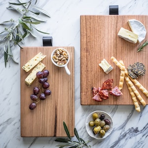 May include: Two wooden serving boards with black leather handles, each laden with food items. One board holds cheese, grapes, and walnuts. The other features cheese, salami, olives, and breadsticks. The boards are on a marble surface.