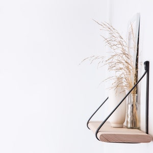 May include: A wooden shelf with black metal brackets, mounted on a white wall. The shelf is decorated with a white vase and a glass vase with dried grasses.