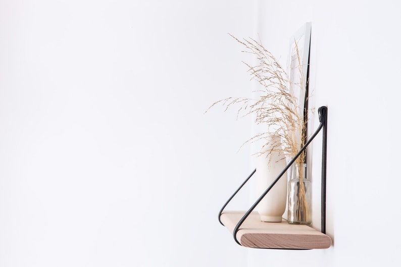 May include: A wooden shelf with black metal brackets, mounted on a white wall. The shelf is decorated with a white vase and a glass vase with dried grasses.
