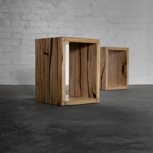 May include: Two open-faced wooden cube-shaped side tables. The tables are made of light brown wood with dark brown grain patterns. The tables are set against a white brick wall and a dark grey floor.