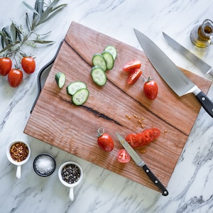 Op de afbeelding: Een houten snijplank met een donkere handgreep, omringd door verse ingrediënten. Gesneden komkommers en tomaten zijn op de plank gerangschikt, samen met twee grote koksmessen en een kleiner schilmes. Kleine bakjes met kruiden en een fles olijfolie maken de scène compleet.