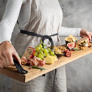 May include: A rectangular wooden serving board laden with cheese, grapes, cured meats, and snacks. The board has black leather handles. The person holding the board is wearing an apron.