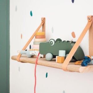May include: A wooden shelf with leather straps hangs on a white wall with colorful dots. The shelf holds a green wooden toy crocodile, a stacking ring toy, and a blue and white toy.