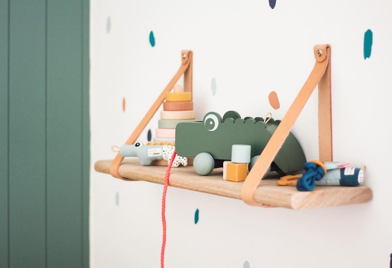 May include: A wooden shelf with leather straps hangs on a white wall with colorful dots. The shelf holds a green wooden toy crocodile, a stacking ring toy, and a blue and white toy.