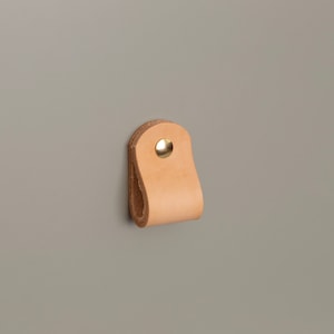 Puede incluir: Un tirador de cajón de cuero de color beige claro con un remache dorado. El tirador en forma de lazo está fijado a una superficie gris. Este diseño minimalista es adecuado para armarios y cajones.