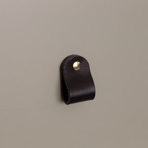 Puede incluir: Un tirador de armario de cuero negro con un remache dorado. El tirador tiene forma de lazo y está fijado a una superficie de color neutro. Esta ferretería es adecuada para cajones, armarios y otros muebles.