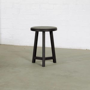 Puede incluir: Un taburete de madera negro con asiento redondo y tres patas anguladas. El taburete tiene un diseño sencillo y minimalista, adecuado para varios estilos de interior. El taburete mide aproximadamente 46 cm de alto.