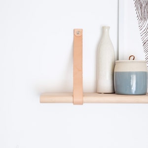 May include: A light wood shelf with a leather strap and two ceramic containers. One container is a tall, white bottle and the other is a shorter, round container with a light blue glaze.