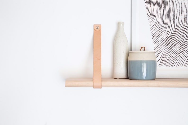May include: A light wood shelf with a leather strap and two ceramic containers. One container is a tall, white bottle and the other is a shorter, round container with a light blue glaze.