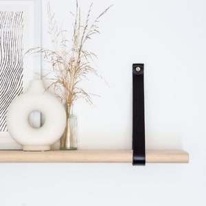 May include: A wooden shelf with a black leather strap attached to the wall. The shelf is decorated with a white vase, dried flowers, and a framed print.