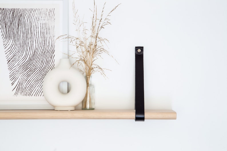 May include: A wooden shelf with a black leather strap attached to the wall. The shelf is decorated with a white vase, dried flowers, and a framed print.