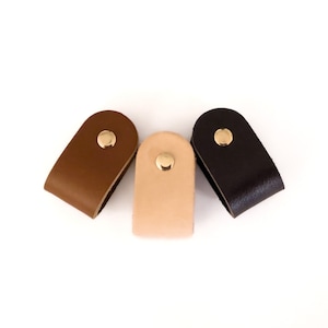 Könnte beinhalten: Drei Leder-Schubladengriffe in Braun, Beige und Schwarz mit goldenen Metallakzenten.