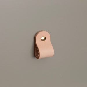 Puede incluir: Un tirador de armario de cuero color beige claro con un remache de latón. El tirador tiene forma de lazo, fijado a una superficie gris. El cuero tiene un aspecto natural, sin terminar.