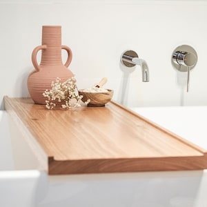 Puede incluir: Una bandeja de bañera de madera clara, con un jarrón de terracota, un pequeño cuenco de madera con una cuchara y flores blancas. La bandeja abarca el ancho de una bañera blanca, con grifos cromados en la pared.