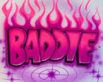 May include: A white fabric surface featuring airbrushed graffiti-style lettering in shades of pink and magenta that spells the word BADDIE. The text is surrounded by stylized pink flames at the top and a circular target graphic with four-pointed stars.