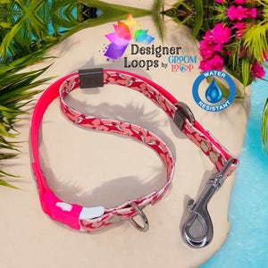 May include: Pink and white dog collar with a shark print and a silver clip. The collar is water resistant and has the text "Designer Loops by Groom Loop" on it.
