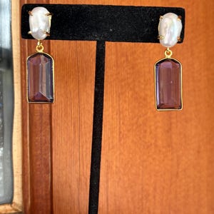 Peut inclure: Une paire de boucles d'oreilles dorées. Chaque boucle d'oreille présente une perle blanche de forme ovale en haut, reliée à une pierre précieuse rectangulaire violette. Les pierres précieuses sont encadrées d'or. Les boucles d'oreilles sont exposées sur un présentoir à bijoux noir.