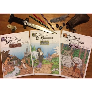 Può includere: Tre libri da colorare con illustrazioni di creature mitiche, favole e bestie. I libri sono intitolati "Colouring Bestiarum" con il numero del volume e una parola descrittiva per ogni libro. I libri sono su una superficie di legno con un pugnale, matite e una penna.