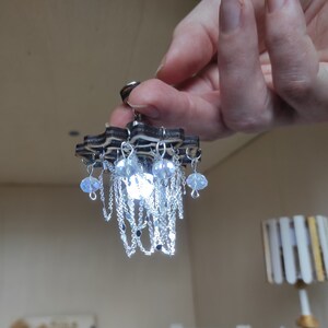 May include: A miniature wooden chandelier with a star-shaped base and crystal-like drops hanging from it. The chandelier is lit up with a white light.