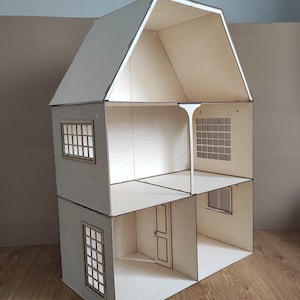 May include: A wooden dollhouse with a white painted finish. The dollhouse has three levels, a pitched roof, and windows with black frames. The dollhouse is sitting on a wooden floor.