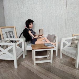 May include: A doll sitting on a white couch with a laptop, a white table, and two white chairs with pillows. The doll has black hair and is wearing a blue and white plaid shirt and blue denim shorts.