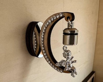 Op de afbeelding: Een houten halve maan met strass steentjes, een zilveren engelbeeldje en een zilveren bel die eraan hangt.